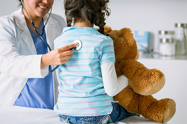 female provider in white coat placing stethoscope on the back of a young girl who is holding a teddy bear