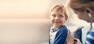 Young blonde boy smiling towards provider who is placing a stethoscope on his back
