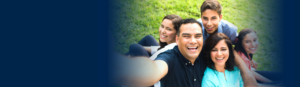 Hispanic family of 4 sitting on a picnic blanket with dad taking a selfie 